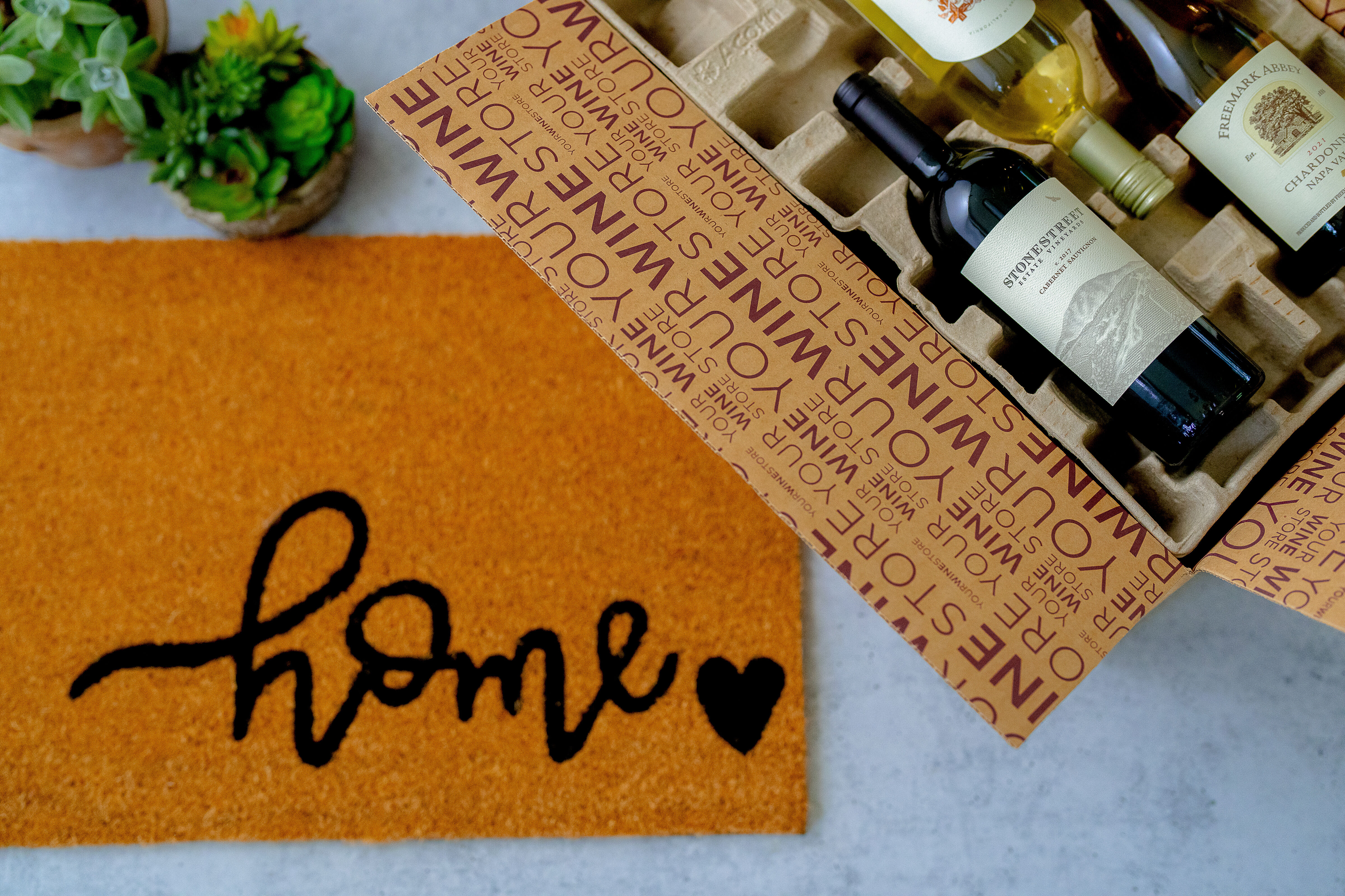 Bottles of wine in a shipping box, on a doorstep with a rug that says 'home'